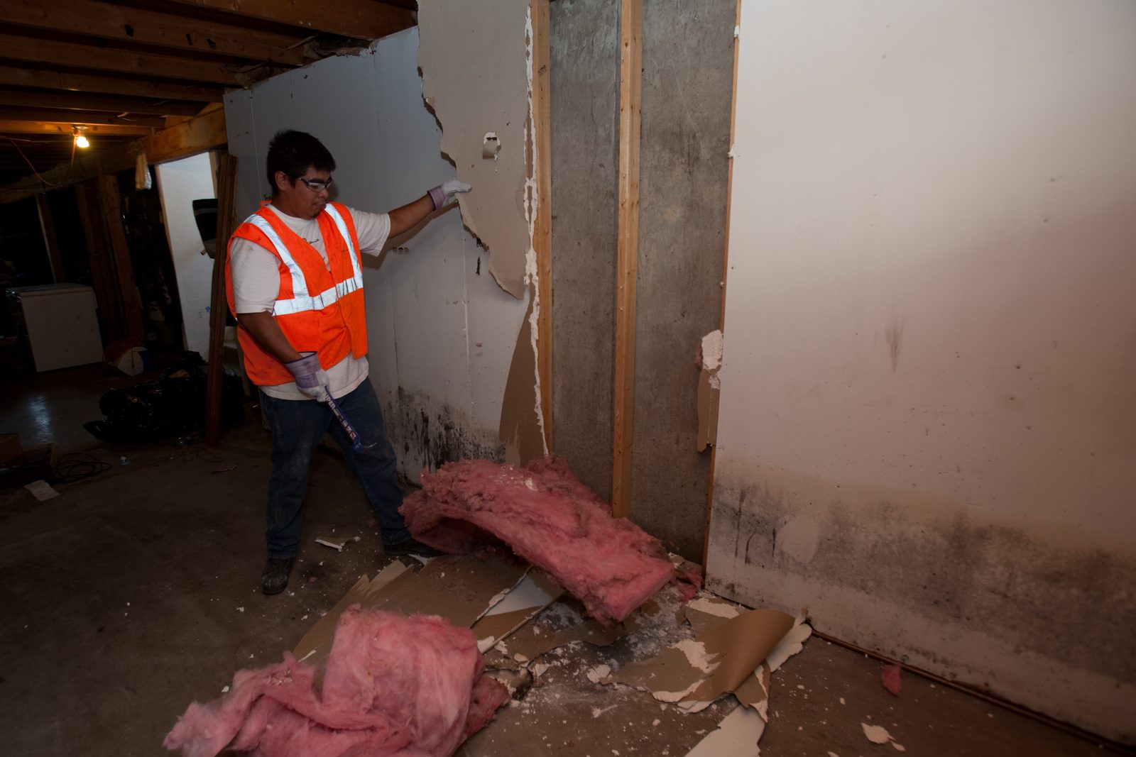 Technician in a high-visibility vest pulling wet drywall and insulation off a basement wall